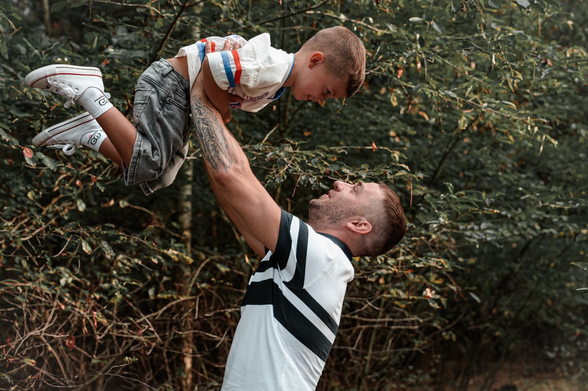 Tata podnosi synka do góry na tle zieleni, widoczne tatuaże na ramieniu ojca i zabawne skarpetki syna z motywem sowy.