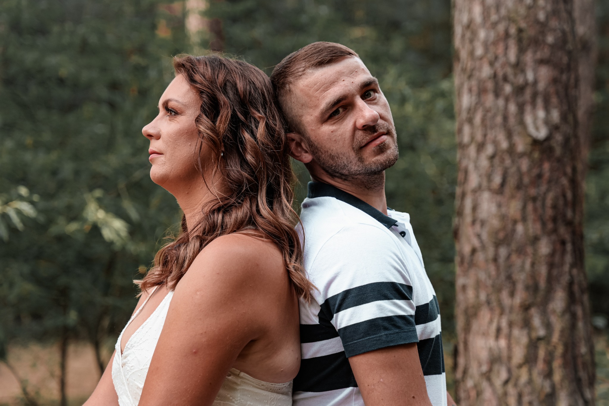 Para opiera się plecami o siebie na tle drzew: kobieta patrzy w lewo, mężczyzna spogląda w obiektyw.