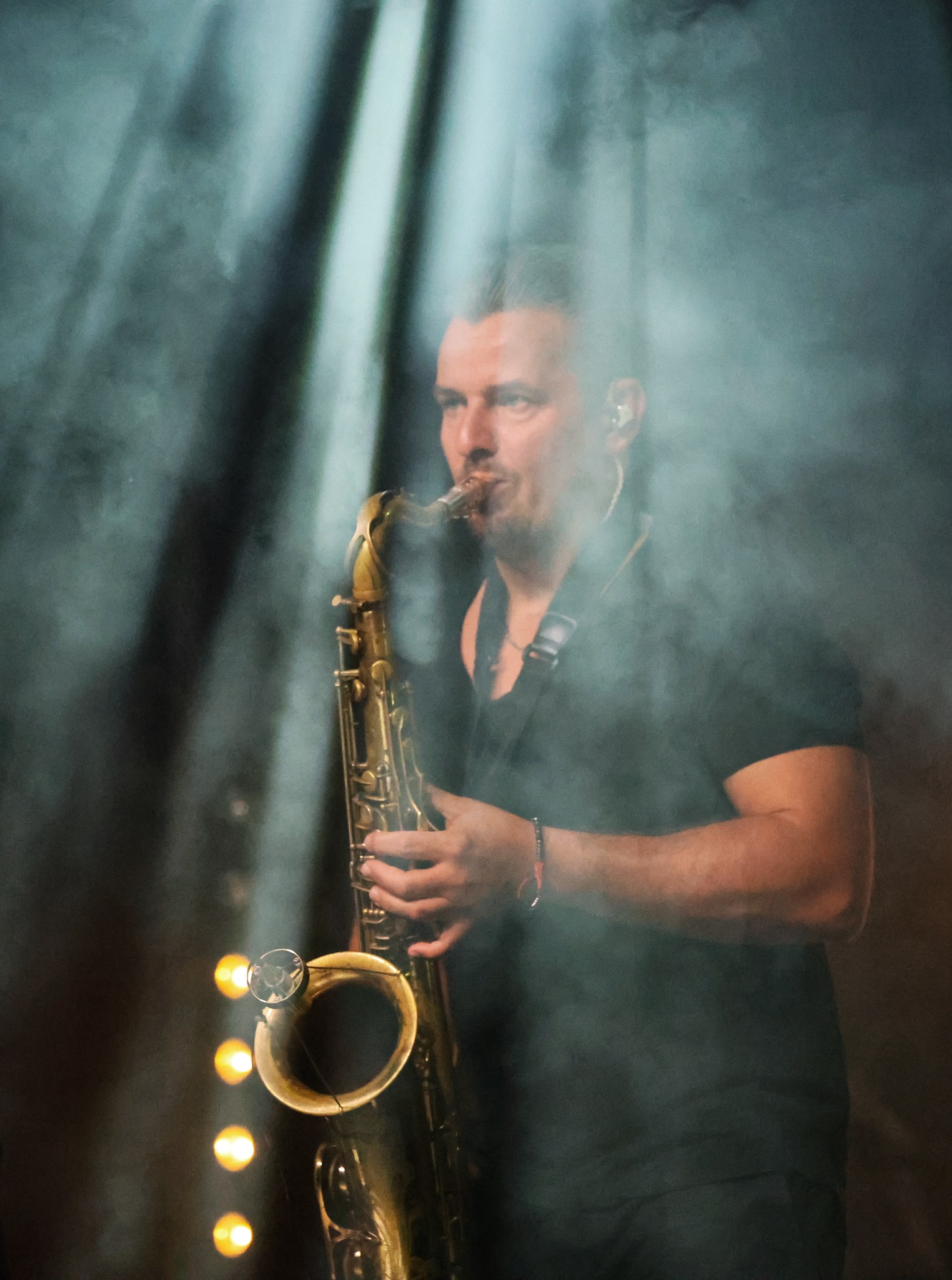 Saksofonista grający na saksofonie tenorowym, oświetlony reflektorami w zadymionym otoczeniu, widoczne słuchawki douszne