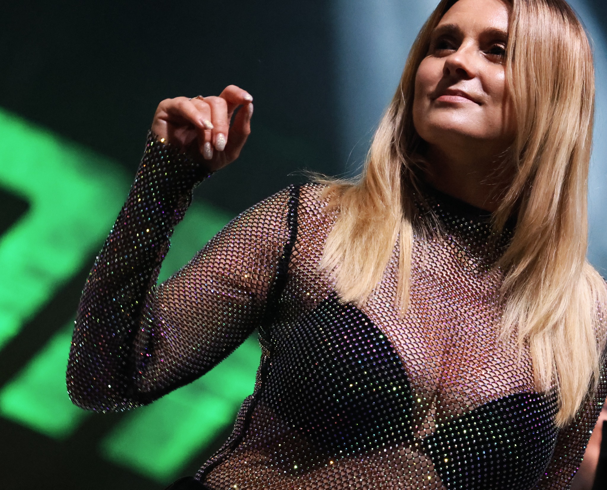Portret kobiety z blond włosami, ubranej w błyszczącą, siatkową bluzkę na scenie z zielonym oświetleniem.