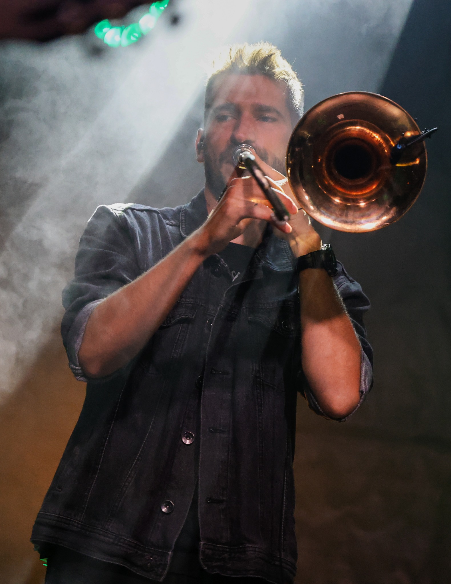 Mężczyzna w dżinsowej kurtce gra na puzonie w zadymionym otoczeniu ze światłami scenicznymi.