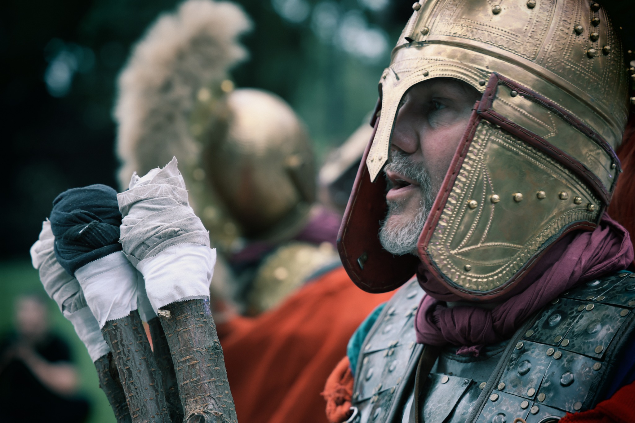 Mężczyzna w historycznym kostiumie rzymskiego legionisty, z metalową zbroją i hełmem, trzymający w ręku trzy patyki owinięte materiałem, z rozmytym tłem przedstawiającym innych ludzi w podobnych...