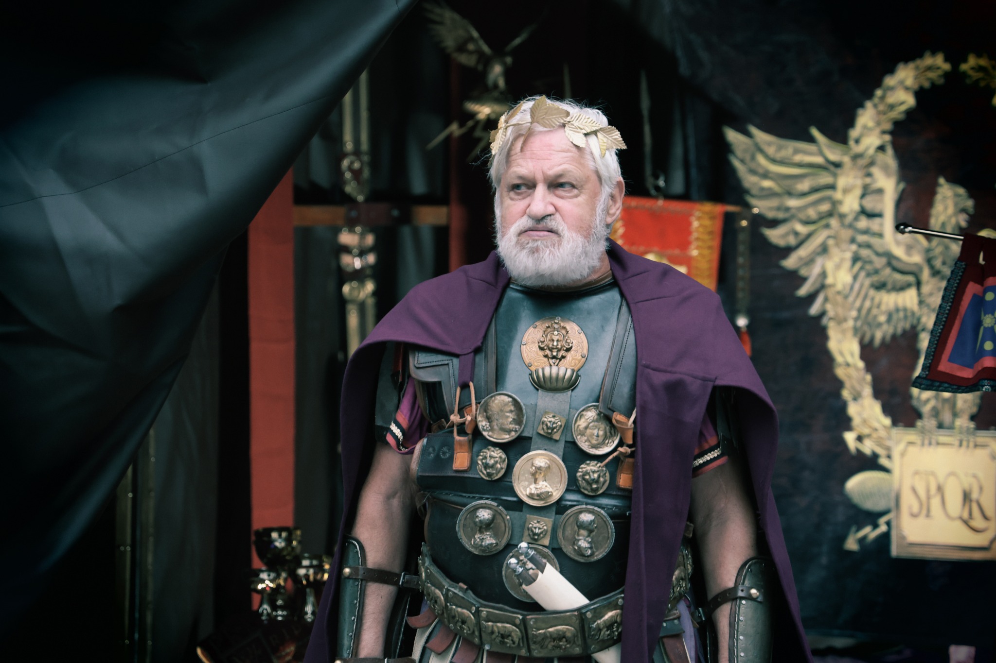 Mężczyzna w kostiumie rzymskiego cesarza z wieńcem laurowym na głowie, w zbroi zdobionej medalionami i purpurowej todze, na tle dekoracji scenicznej z motywem orła i napisem SPQR.