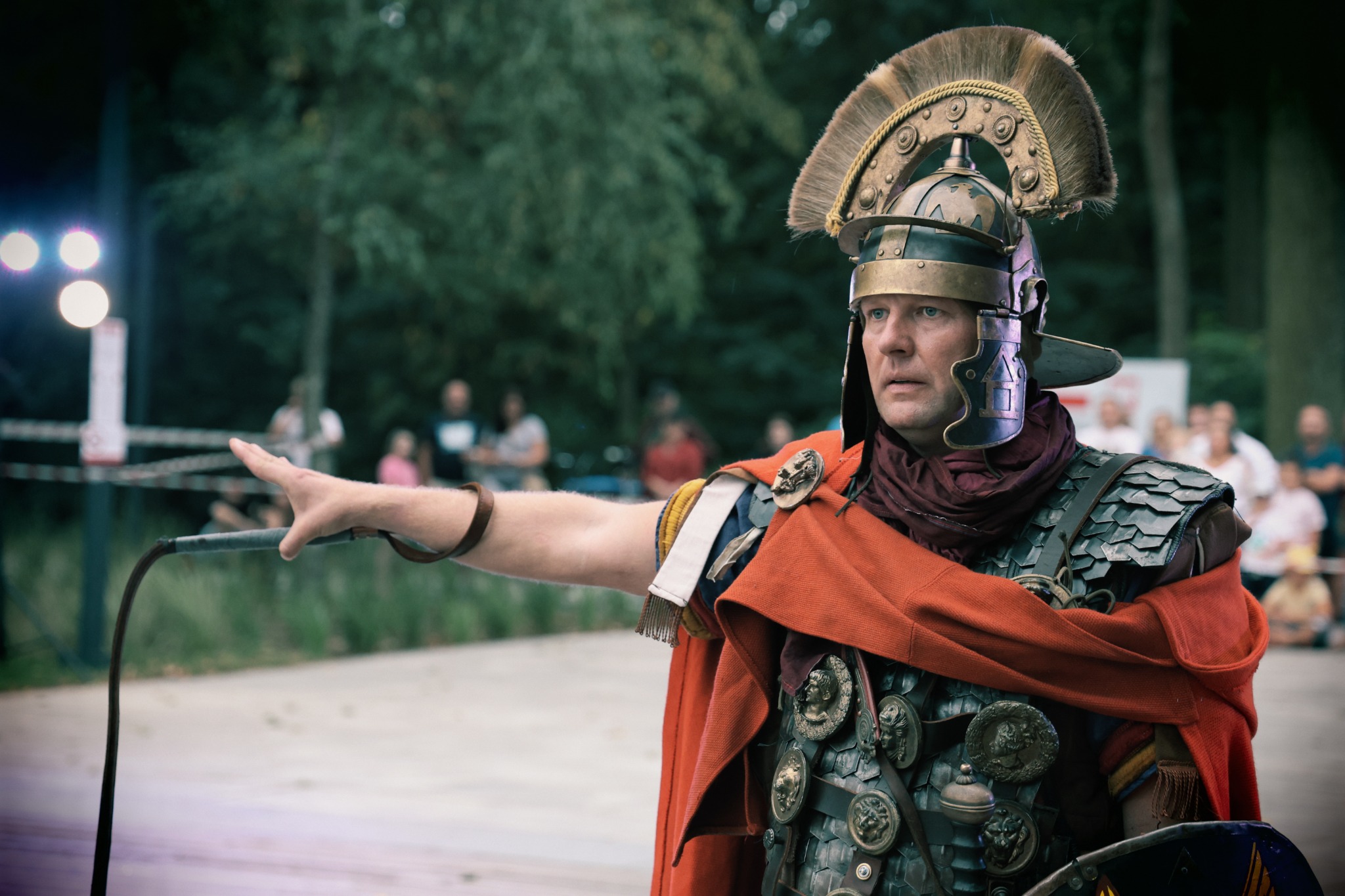 Mężczyzna przebrany za rzymskiego legionistę w pełnym rynsztunku, w hełmie z pióropuszem, z batem w ręku, wskazujący palcem w dal, na tle zieleni i obserwujących go ludzi.