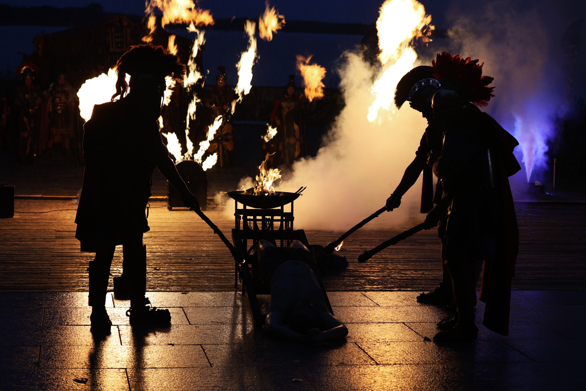 Mroczne widowisko plenerowe: sylwetki aktorów w kostiumach rzymskich z płonącymi pochodniami i ofiarą na katafalku w tle ognia i dymu.