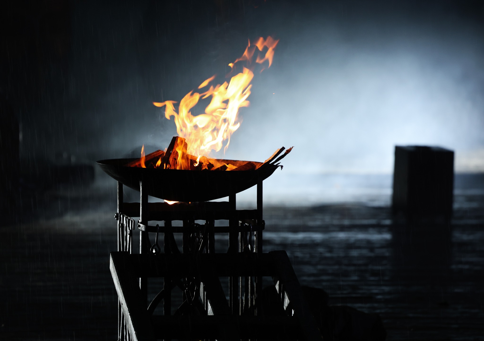Płonące ognisko w metalowej misie na ozdobnym stojaku, padający deszcz, ciemne tło.