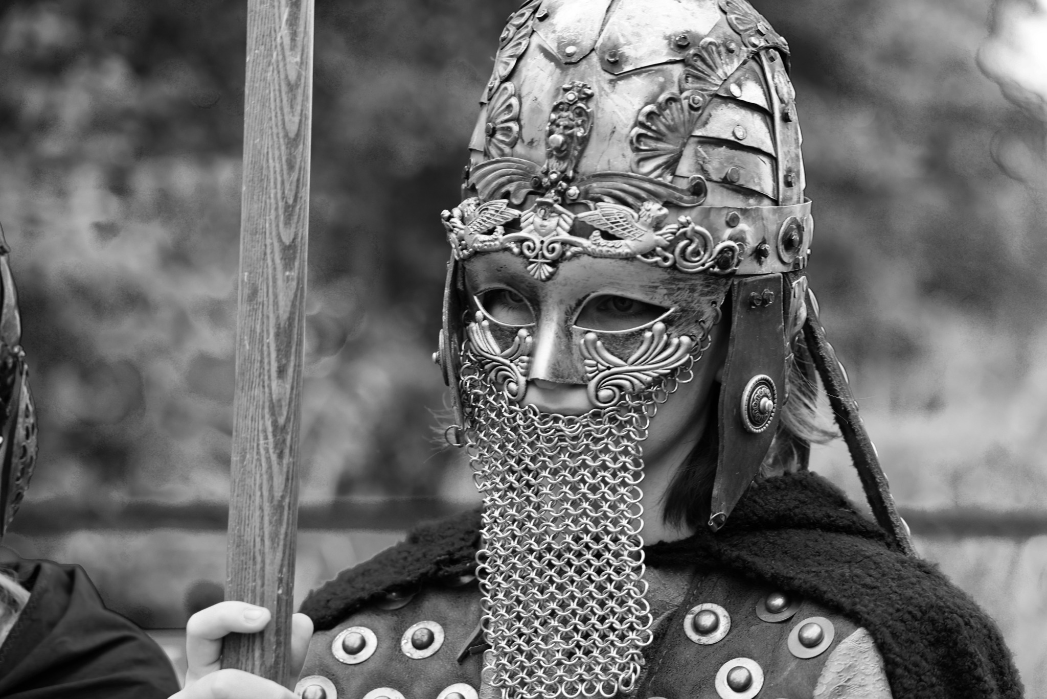 Czarno-białe zbliżenie osoby w bogato zdobionym hełmie i zbroi, z maską na twarzy i kolczugą zakrywającą usta, trzymającej drewniany kij.