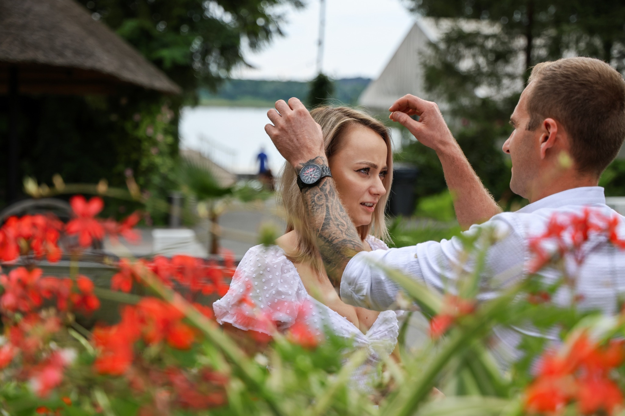 Kobieta z tatuażem na ramieniu i zegarkiem na ręce, mężczyzna poprawia jej włosy na tle jeziora i kwiatów.