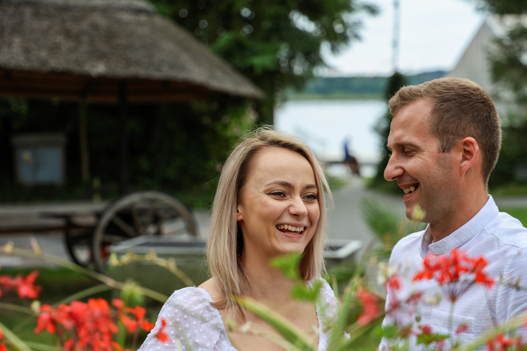 Uśmiechnięta para w plenerze, kobieta blondynka w białej sukience, mężczyzna w białej koszuli, w tle altana z trzciny i jezioro.