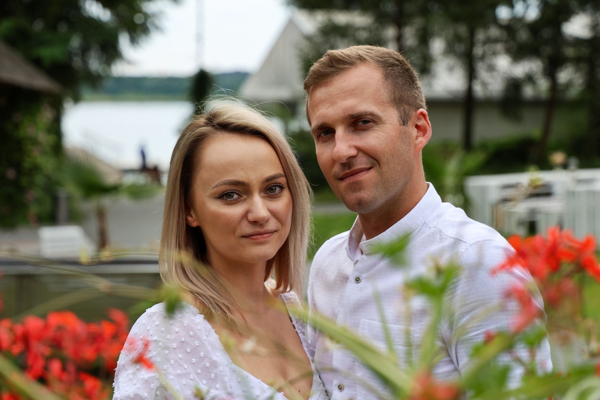 Portret pary w białych strojach na tle zieleni i czerwonych kwiatów, widoczny fragment jeziora i budynek w oddali.