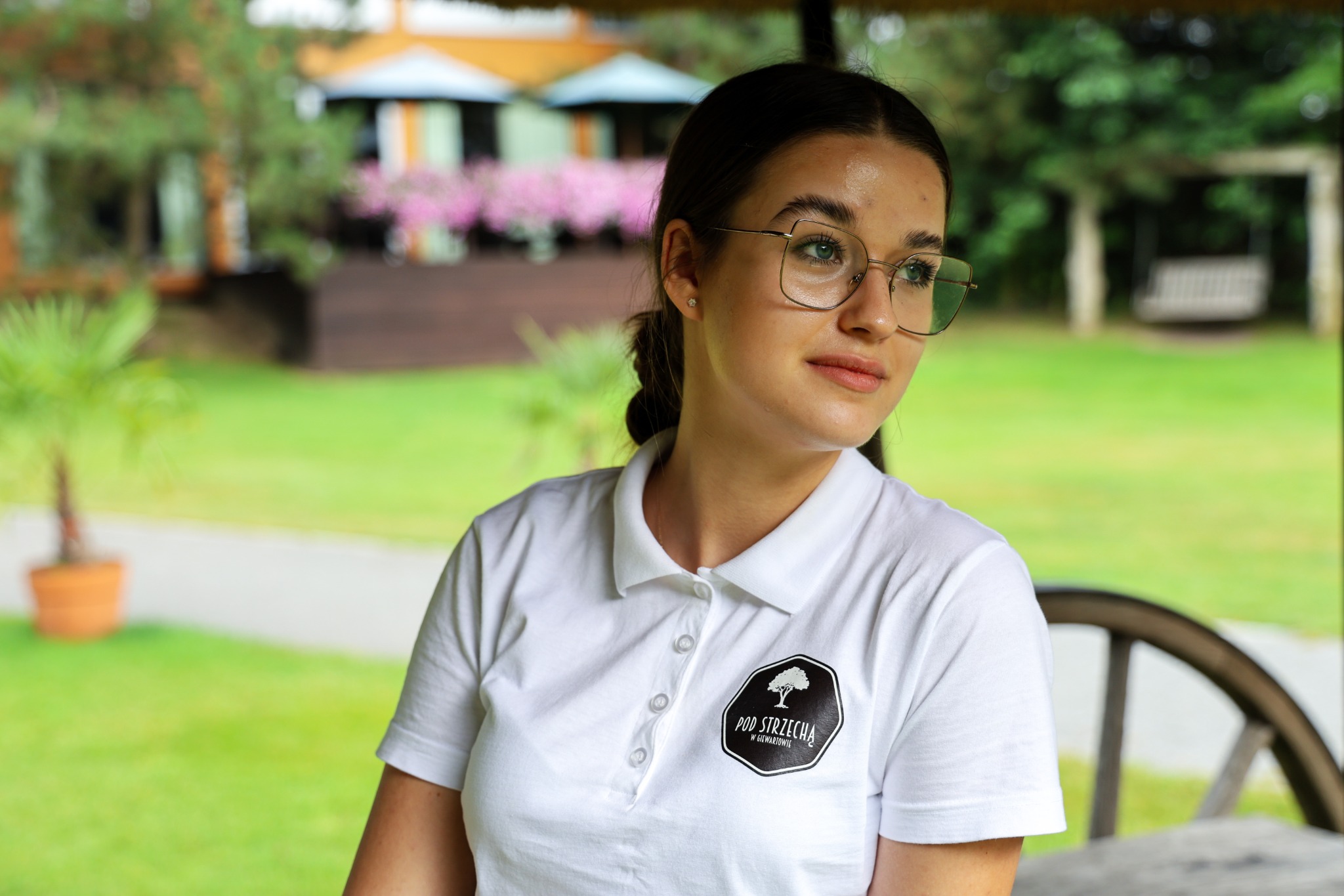 Portret kobiety w okularach, ubrana w białą koszulkę polo z logo, w plenerze z zielenią i budynkami w tle.