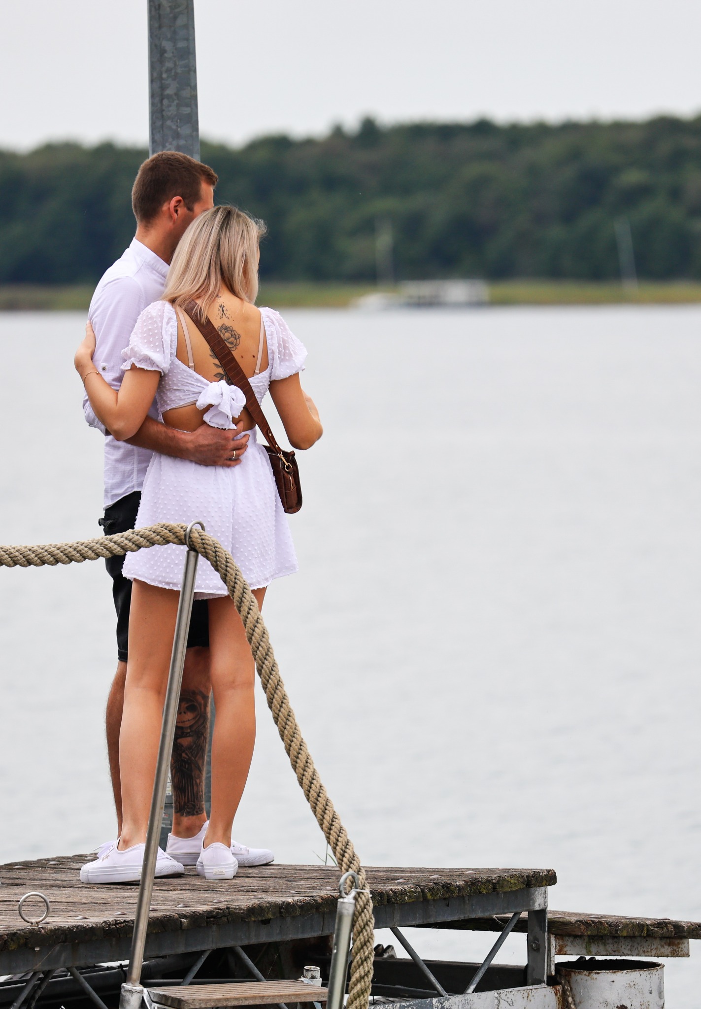 Para obejmująca się na drewnianym pomoście nad wodą, kobieta w białej sukience z odkrytymi plecami i tatuażem kwiatowym, mężczyzna w białej koszuli, widok z tyłu, spokojny krajobraz w tle.