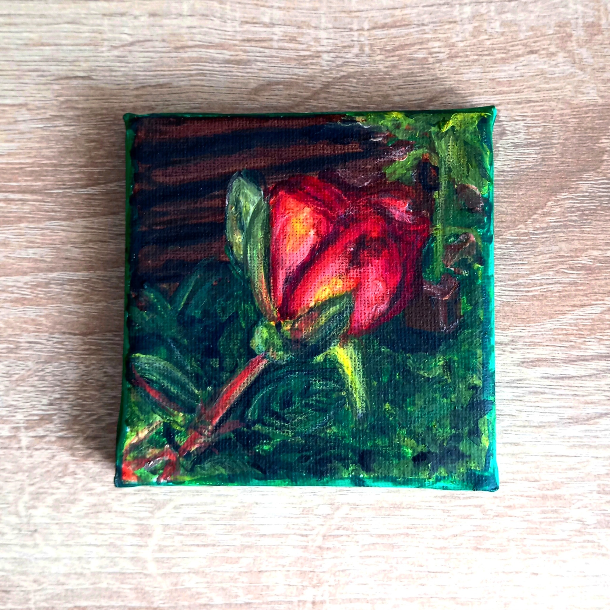 Mały kwadratowy obraz olejny przedstawiający pąk czerwonej róży z zielonymi liśćmi na drewnianym tle.