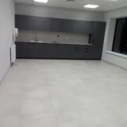 Nowoczesna, szara zabudowa kuchenna w biurowej przestrzeni z jasnymi płytkami podłogowymi i białymi ścianami, widoczny dozownik mydła i grzejnik.