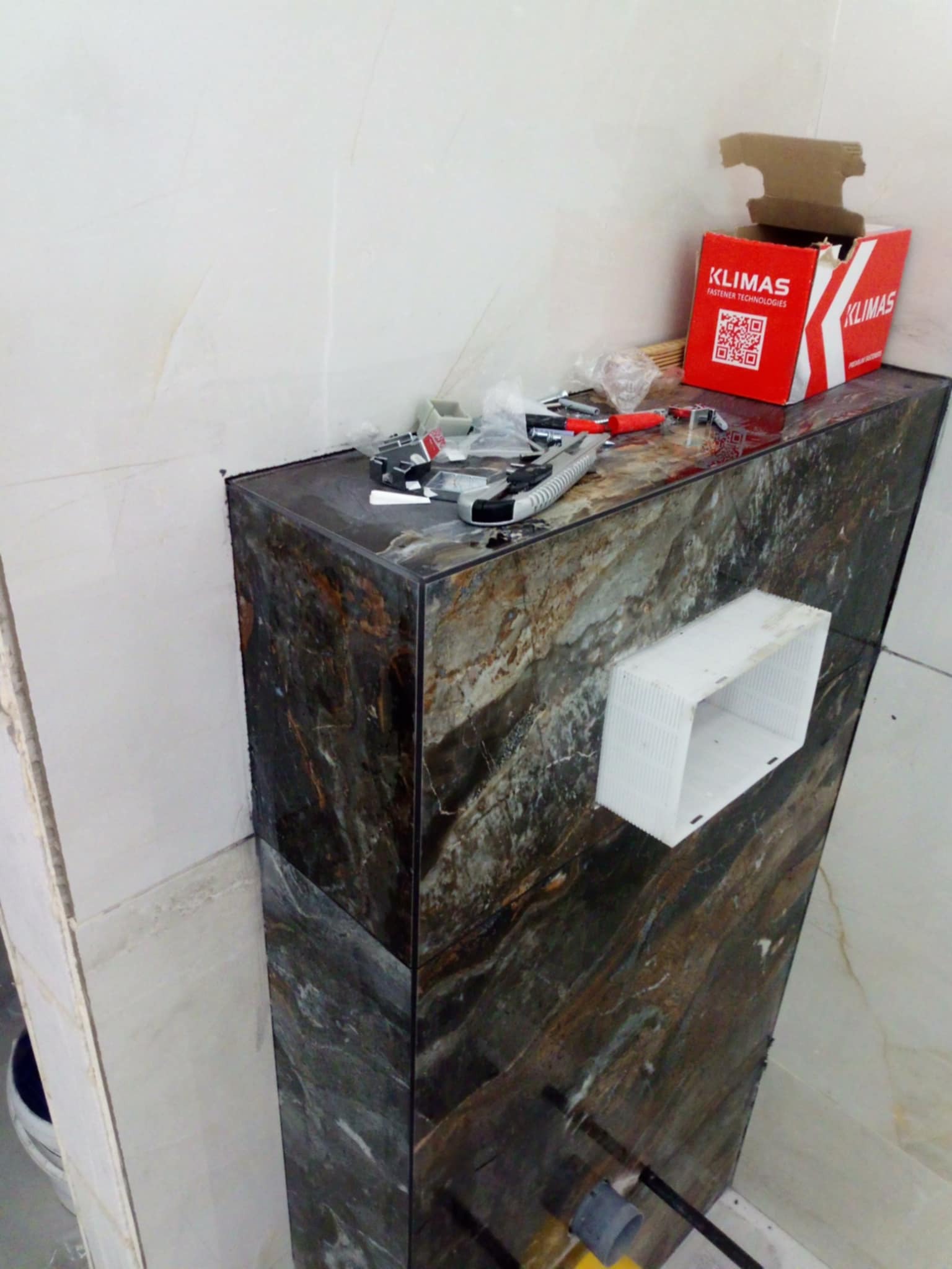 Obudowa stelaża WC wykończona płytkami imitującymi marmur, z widocznym otworem na przyłącze i pudełkiem Klimas na górze, w trakcie prac wykończeniowych.