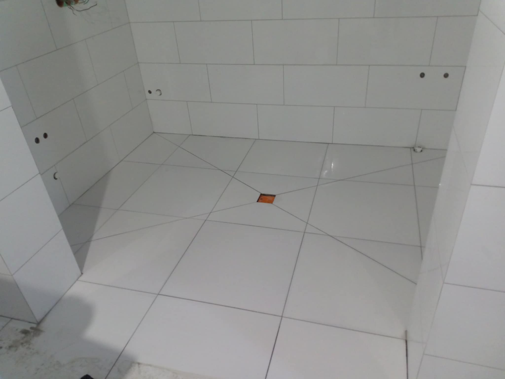 Białe, prostokątne płytki ułożone na ścianach i podłodze w łazience, z widocznym odpływem liniowym w podłodze i otworami na armaturę na ścianie.