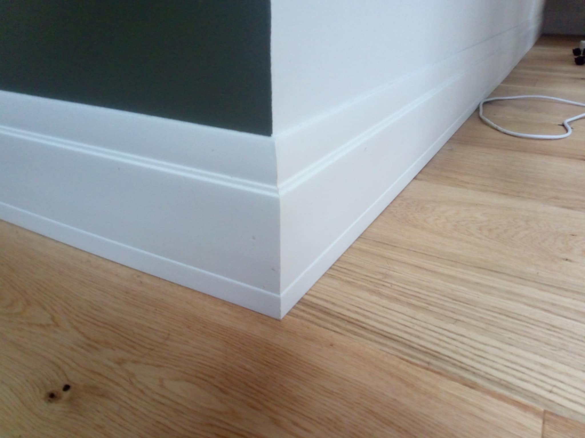 Detal narożnika pokoju z drewnianą podłogą i białą listwą przypodłogową, ściana pomalowana na ciemnozielono i biało