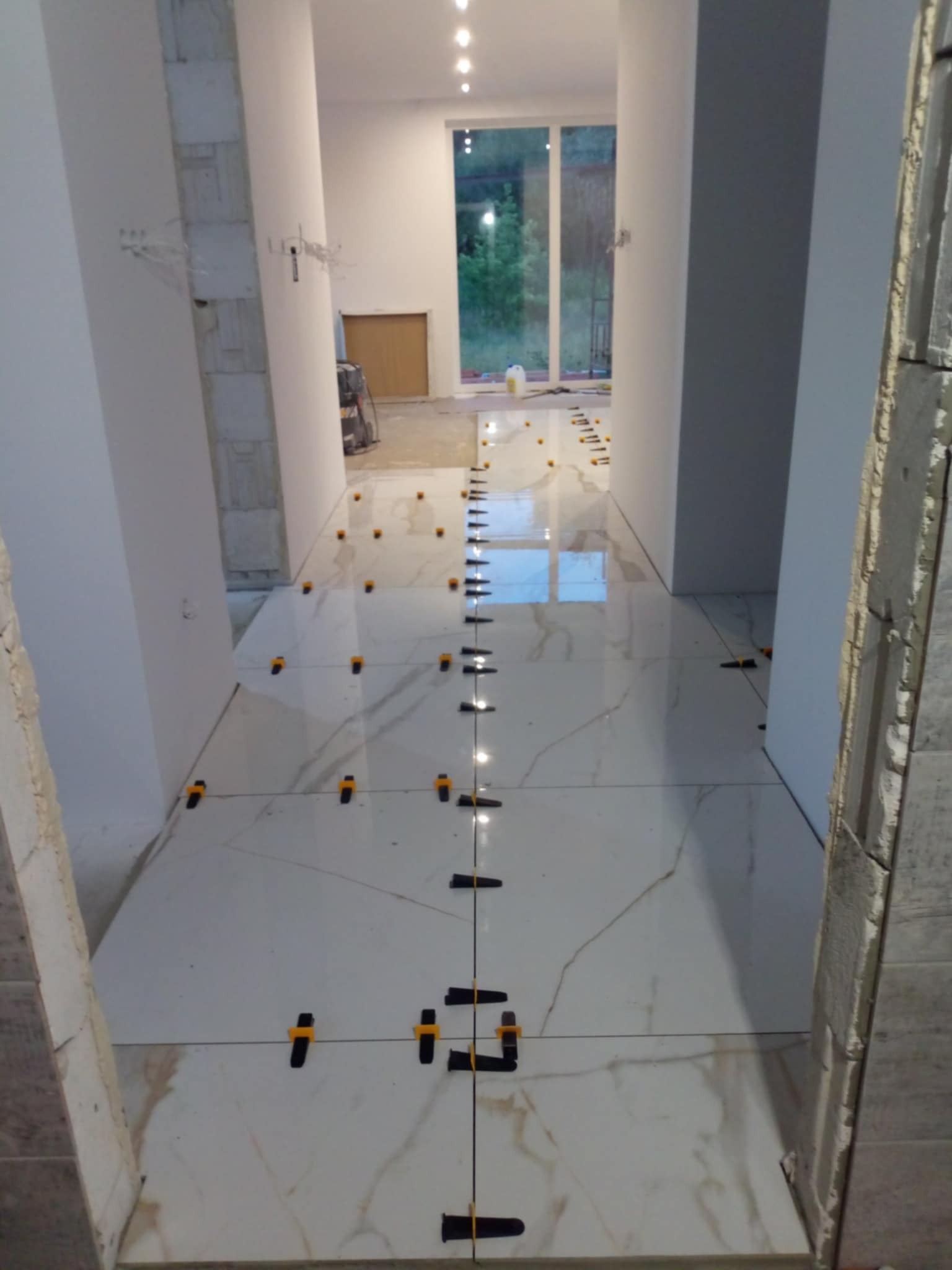 Korytarz w trakcie układania białych płytek imitujących marmur, z użyciem czarnych i żółtych klipsów poziomujących, widoczne niedokończone obłożenie filaru płytkami.