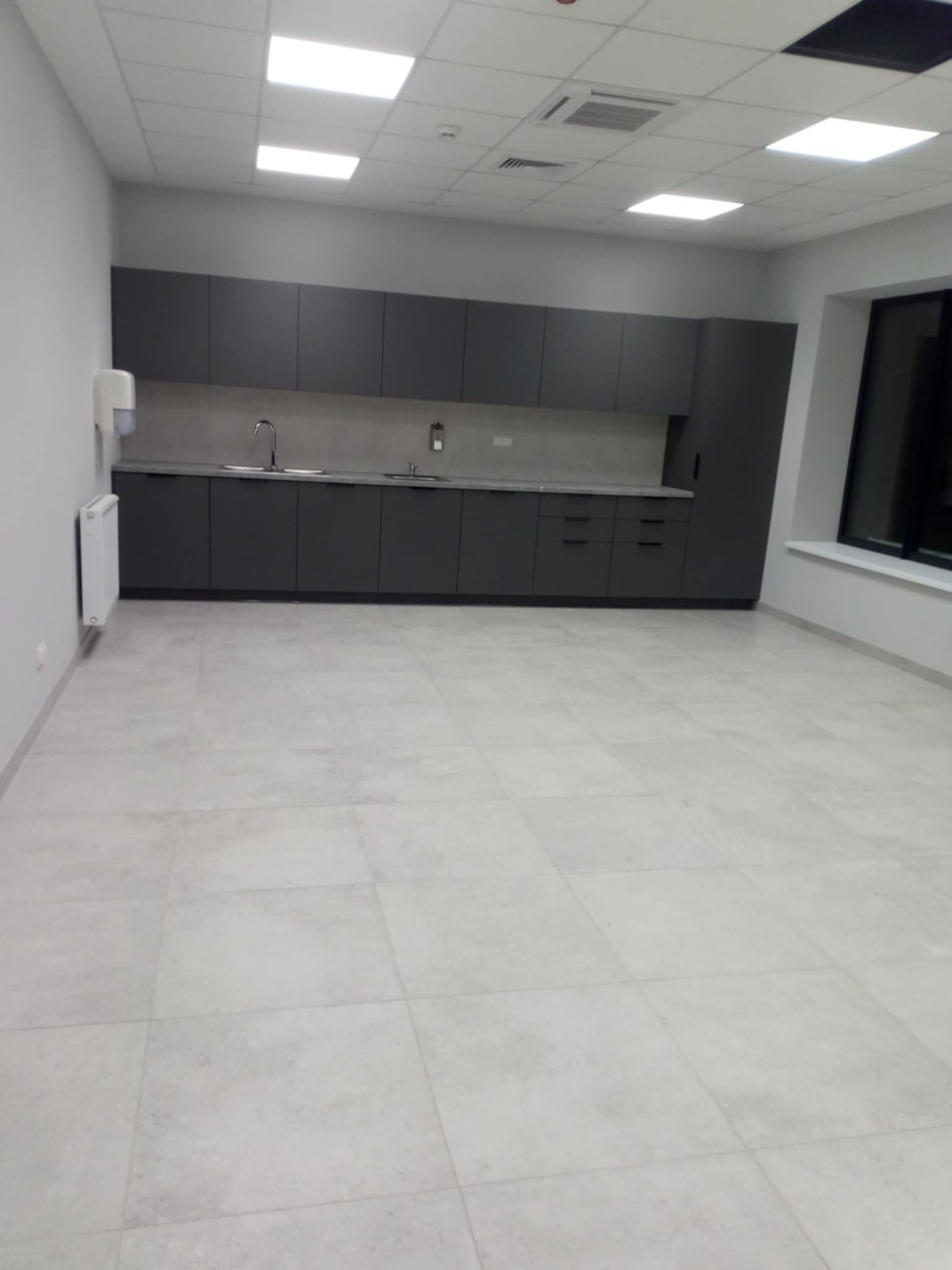 Nowoczesna, szara zabudowa kuchenna w biurowej przestrzeni z jasnymi płytkami podłogowymi i białymi ścianami, widoczny dozownik mydła i grzejnik.