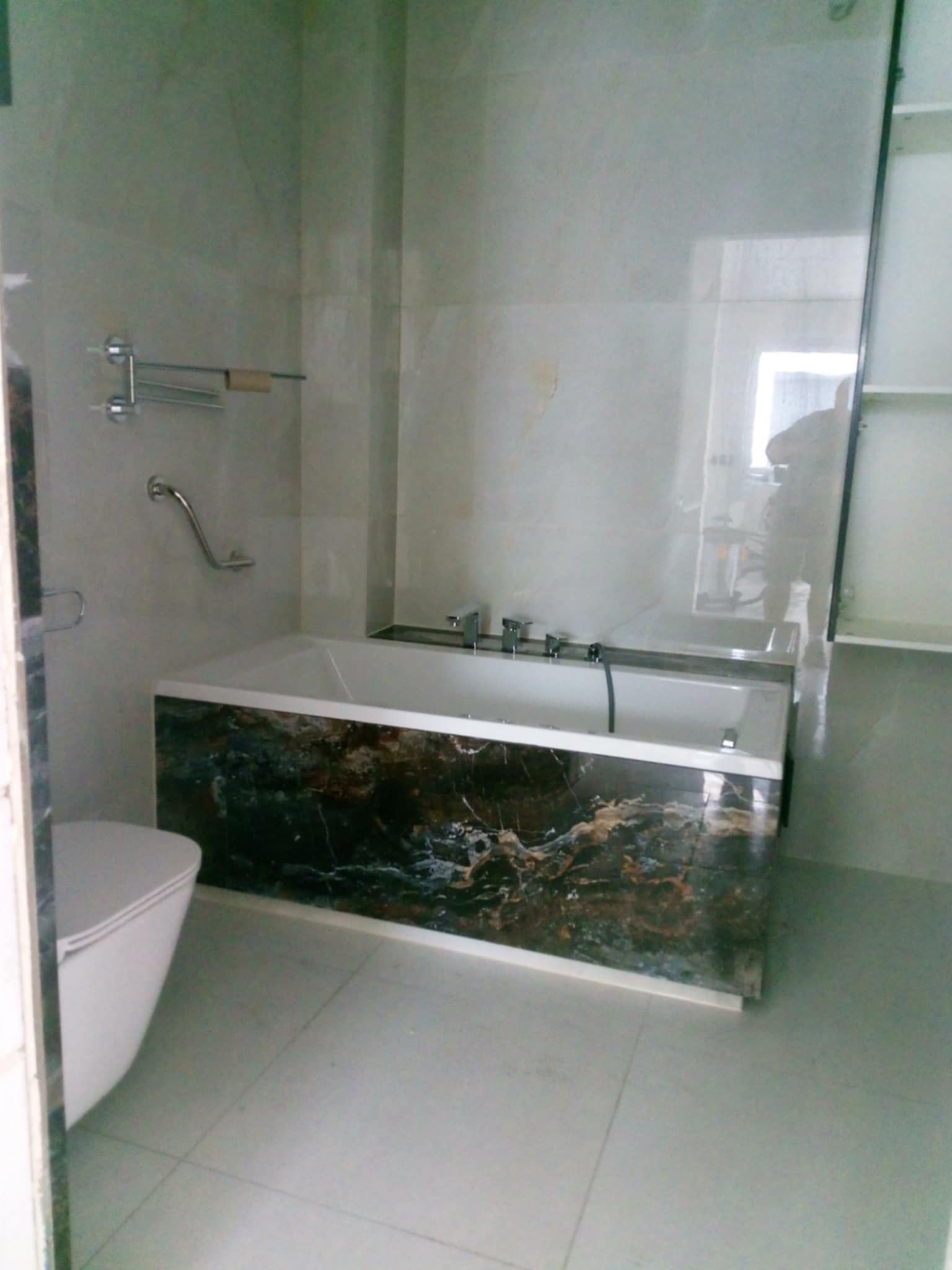 Nowoczesna łazienka z wanną obudowaną marmurem, biała toaleta i szklana kabina prysznicowa, widoczne chromowane akcesoria łazienkowe.