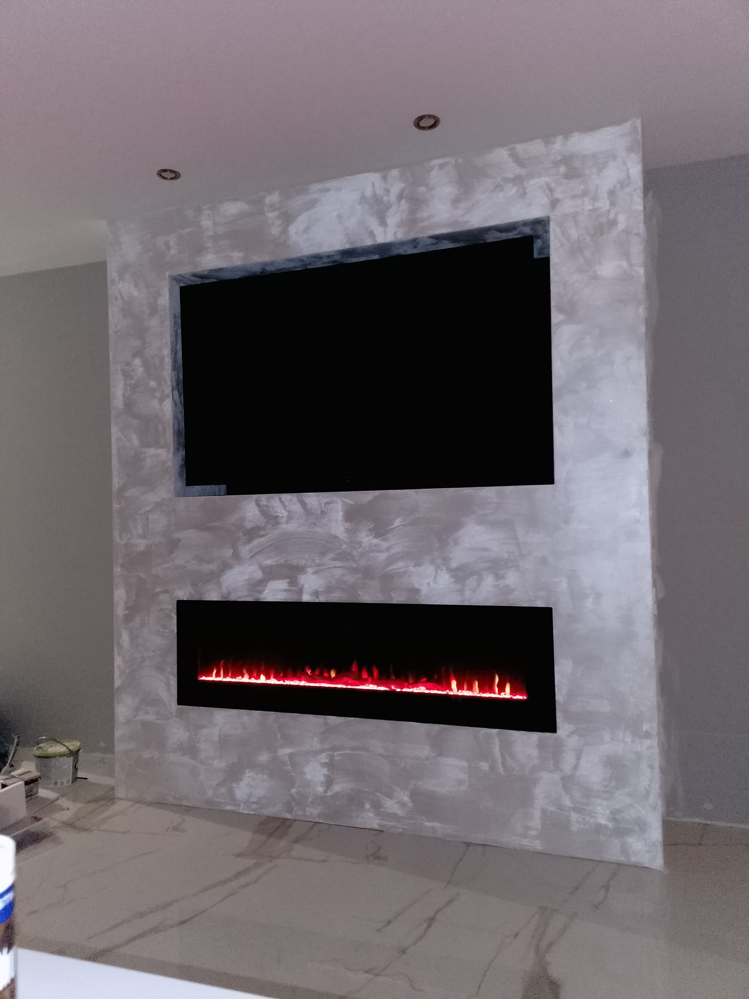 Ściana z dekoracyjnym tynkiem w odcieniach szarości, z wnęką na telewizor i elektrycznym kominkiem. Podłoga wyłożona płytkami imitującymi marmur.