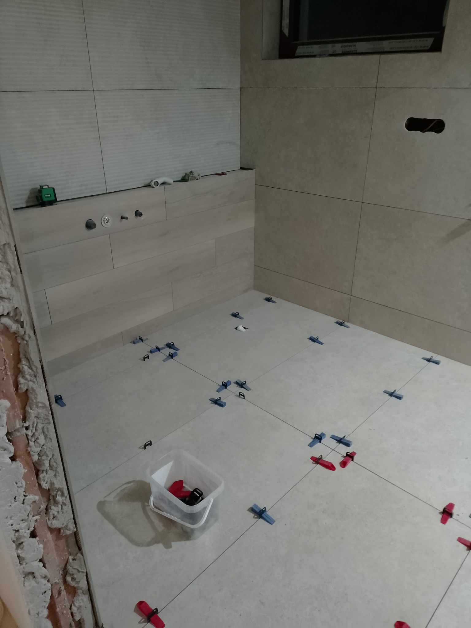 Układanie płytek podłogowych w łazience z użyciem systemu poziomowania. Widoczne krzyżyki dystansowe i pojemnik z klinami. Ściany częściowo wykończone płytkami.