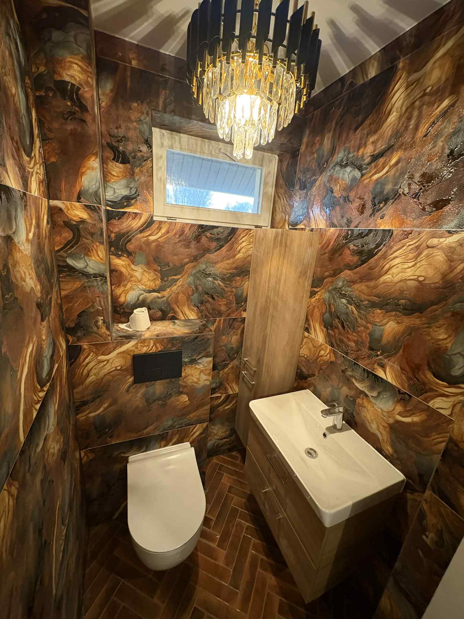Stylowa łazienka z brązowymi płytkami imitującymi marmur, wisząca toaleta, umywalka z szafką i dekoracyjny żyrandol. Nietypowe wykończenie podłogi w jodełkę.
