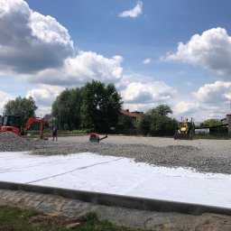 DIGGING RAFAŁ KRZYSZTOŃ - Przygotowanie podłoża pod drogę: rozłożona biała geowłóknina, na niej warstwa szarego kruszywa, w tle pomarańczowa minikoparka i żółta ładowarka, pracownicy w trakcie robót.