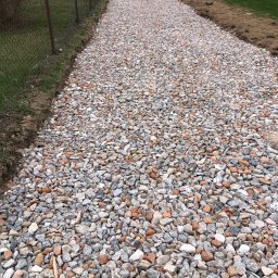 DIGGING RAFAŁ KRZYSZTOŃ - Świeżo utwardzona droga z mieszanki gruzu i kamienia, widoczne dwie małe koparki na dalszym planie, ogrodzenie z siatki po lewej stronie i pole po prawej