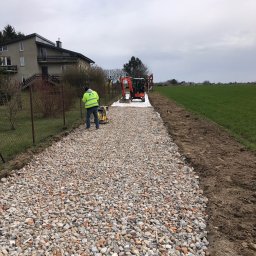 DIGGING RAFAŁ KRZYSZTOŃ - Utwardzanie drogi: Pracownik w żółtej kamizelce zagęszcza gruz na przygotowanym podłożu, w tle minikoparki na białej geowłókninie i zielone pole.
