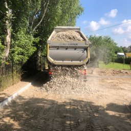 DIGGING RAFAŁ KRZYSZTOŃ - Wywrotka z otwartą skrzynią wysypuje żwir na przygotowany teren z obrzeżami. Widoczne ślady opon i zieleń w tle. Etap utwardzania drogi dojazdowej.