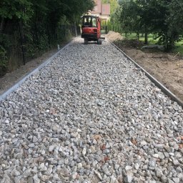 DIGGING RAFAŁ KRZYSZTOŃ - Utwardzona droga z szarym kamieniem i obrzeżami, w tle mała koparka Kubota i zieleń drzew. Widok z perspektywy.