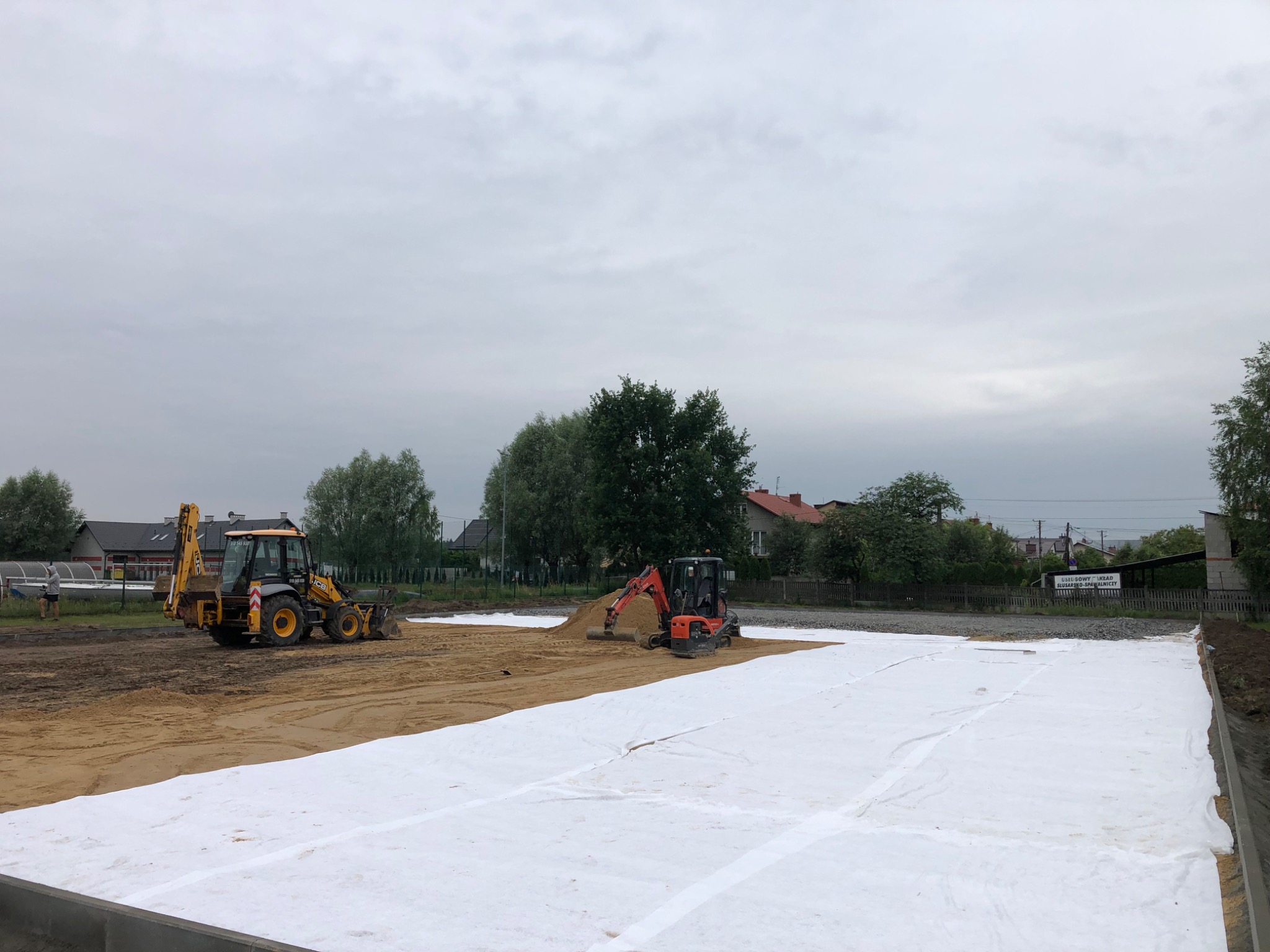 Żółta koparko-ładowarka i pomarańczowa minikoparka na placu budowy z rozłożoną białą geowłókniną, przygotowującym podłoże pod drogę lub parking na tle zabudowań mieszkalnych i pochmurnego nieba.