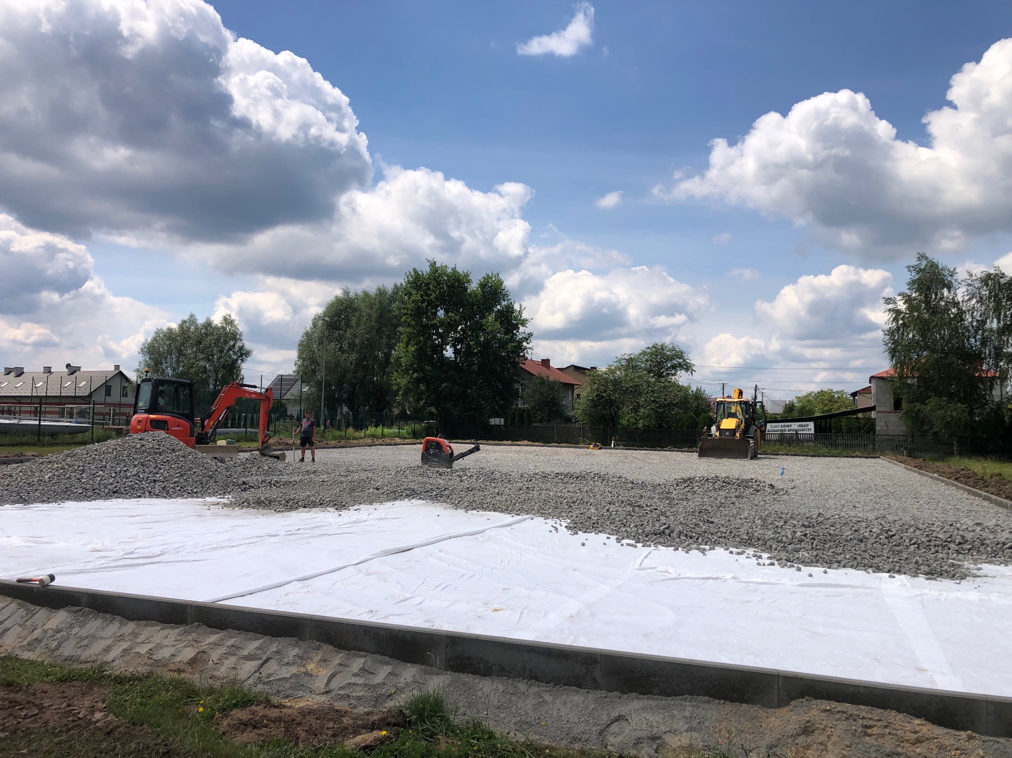Przygotowanie podłoża pod drogę: rozłożona biała geowłóknina, na niej warstwa szarego kruszywa, w tle pomarańczowa minikoparka i żółta ładowarka, pracownicy w trakcie robót.