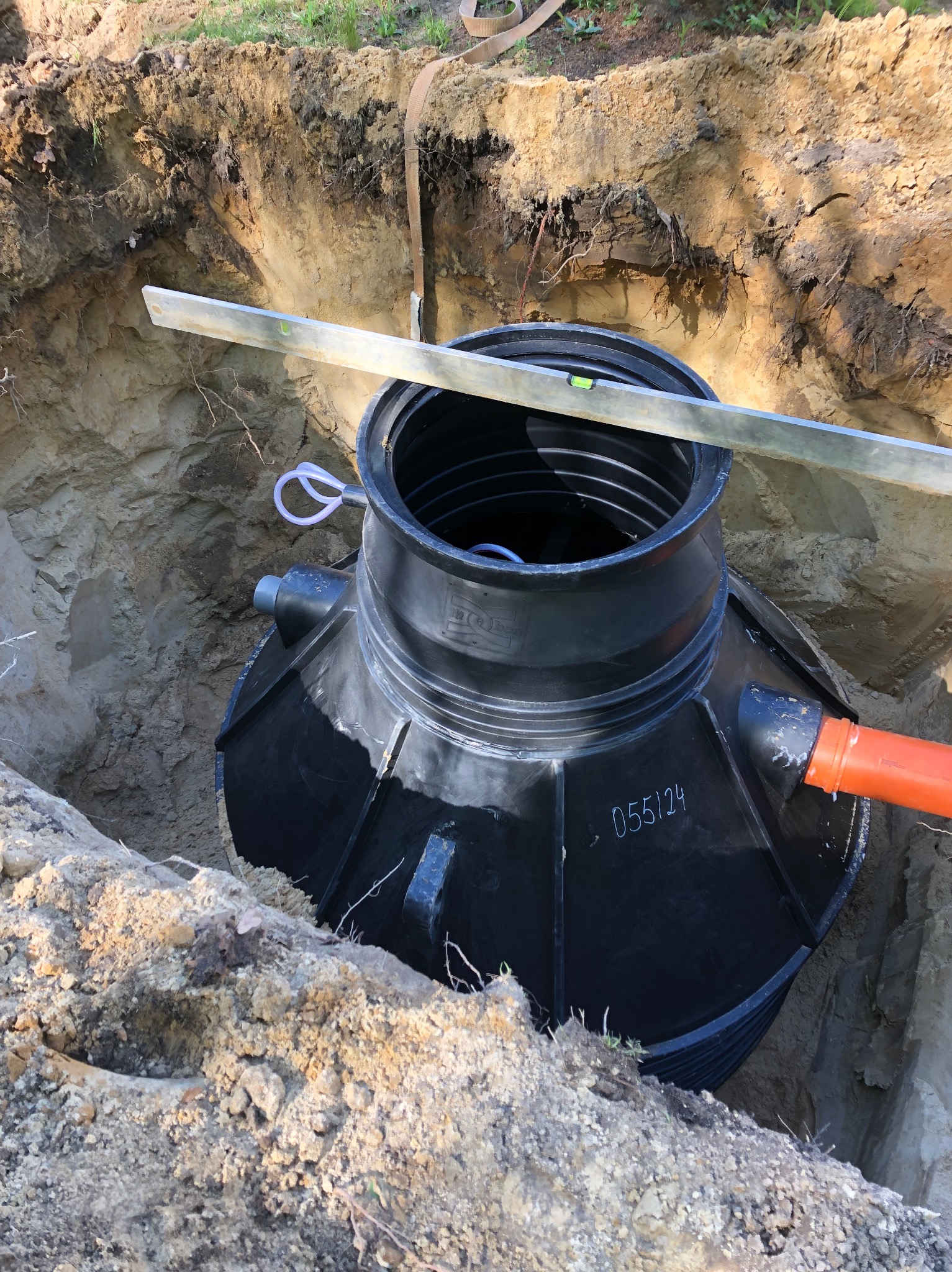 Czarny zbiornik z tworzywa sztucznego umieszczony w wykopie, poziomowany przy użyciu poziomicy, widoczny pomarańczowy przewód odpływowy.
