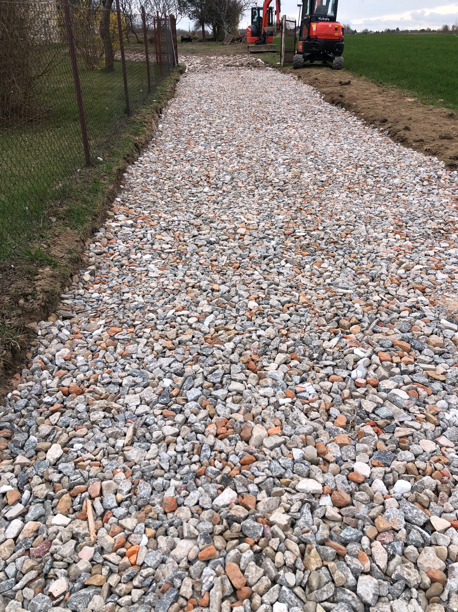 Świeżo utwardzona droga z mieszanki gruzu i kamienia, widoczne dwie małe koparki na dalszym planie, ogrodzenie z siatki po lewej stronie i pole po prawej
