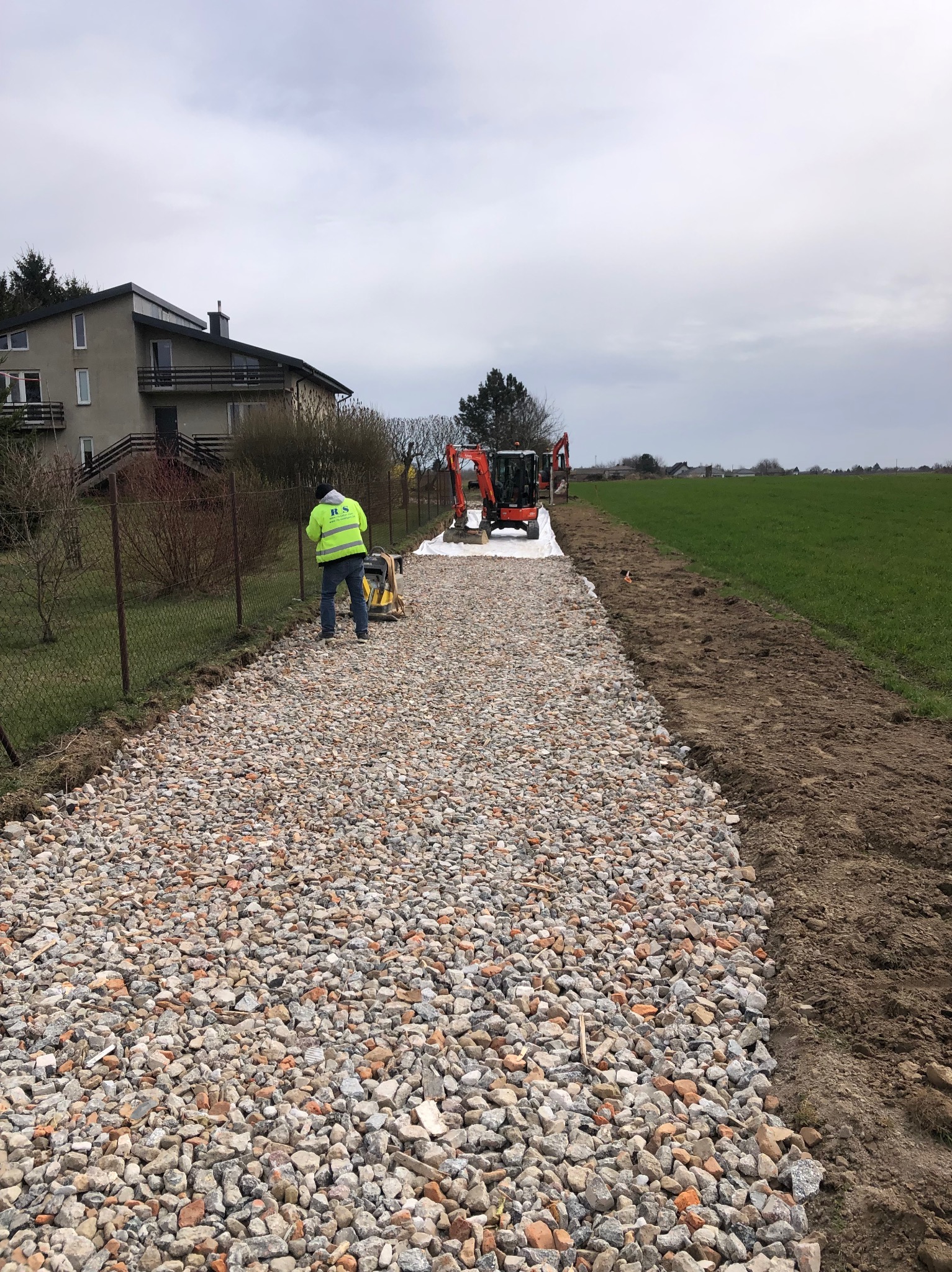 Utwardzanie drogi: Pracownik w żółtej kamizelce zagęszcza gruz na przygotowanym podłożu, w tle minikoparki na białej geowłókninie i zielone pole.