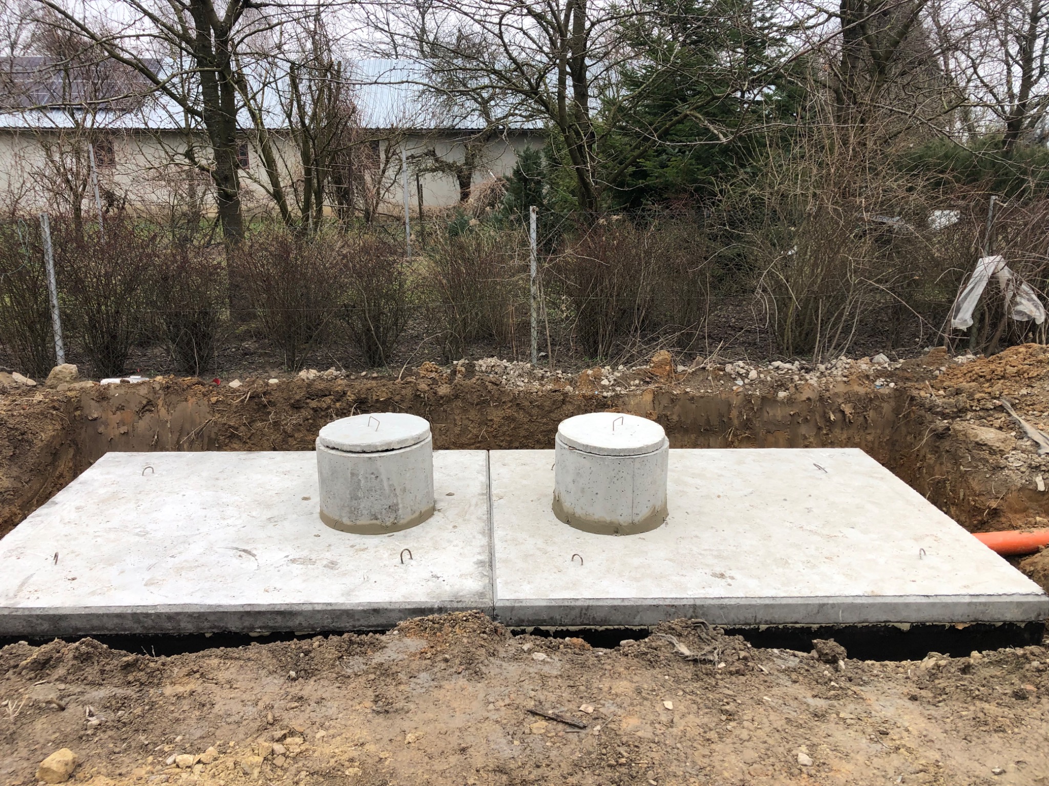 Dwa betonowe zbiorniki z pokrywami w wykopie, widoczna pomarańczowa rura, w tle zabudowania gospodarcze i drzewa bez liści.