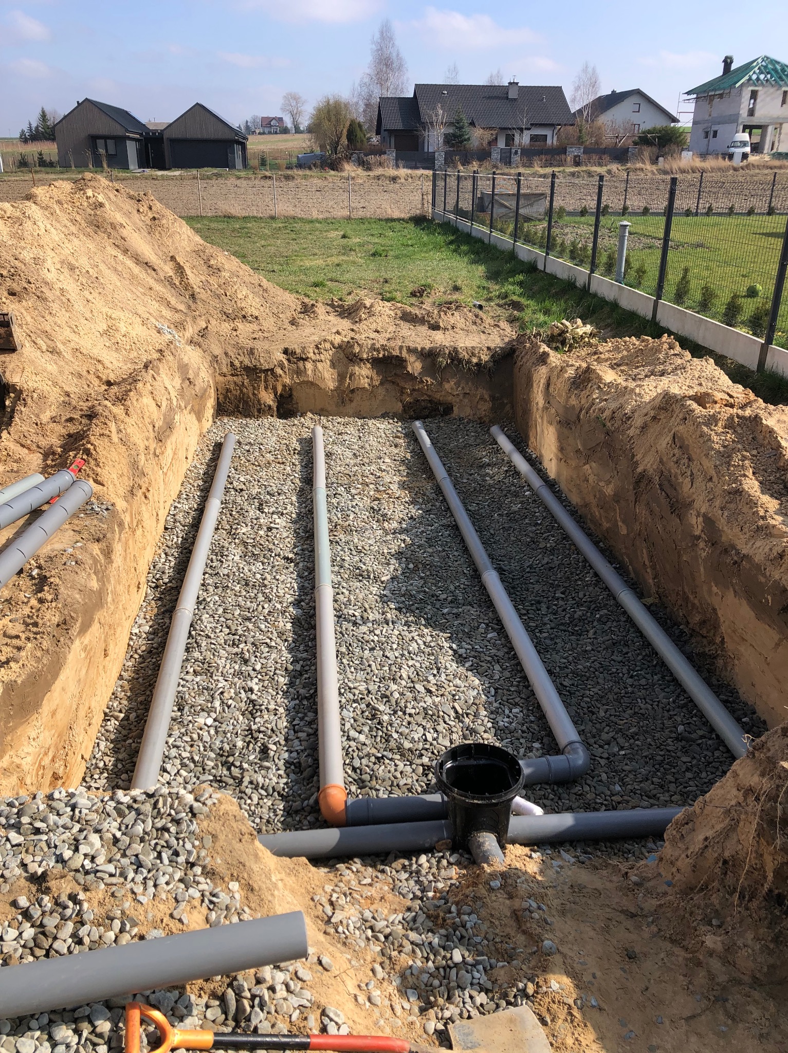 Wykop w ziemi wypełniony żwirem, w którym ułożone są szare rury kanalizacyjne z elementami łączącymi, widoczne narzędzia i fragmenty ziemi, w tle zabudowa jednorodzinna i ogrodzenie.