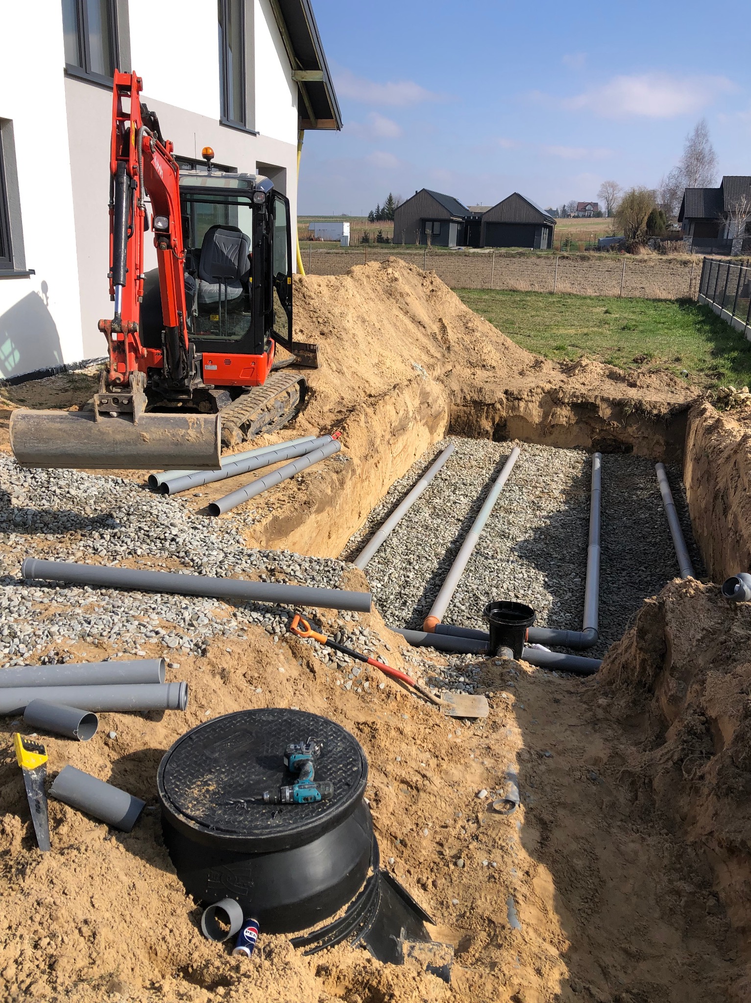 Wykonany wykop pod system rur drenarskich z warstwą żwiru, widoczny minikoparka, czarna studzienka rewizyjna z wiertarką akumulatorową, oraz narzędzia do cięcia rur na tle domu jednorodzinnego...