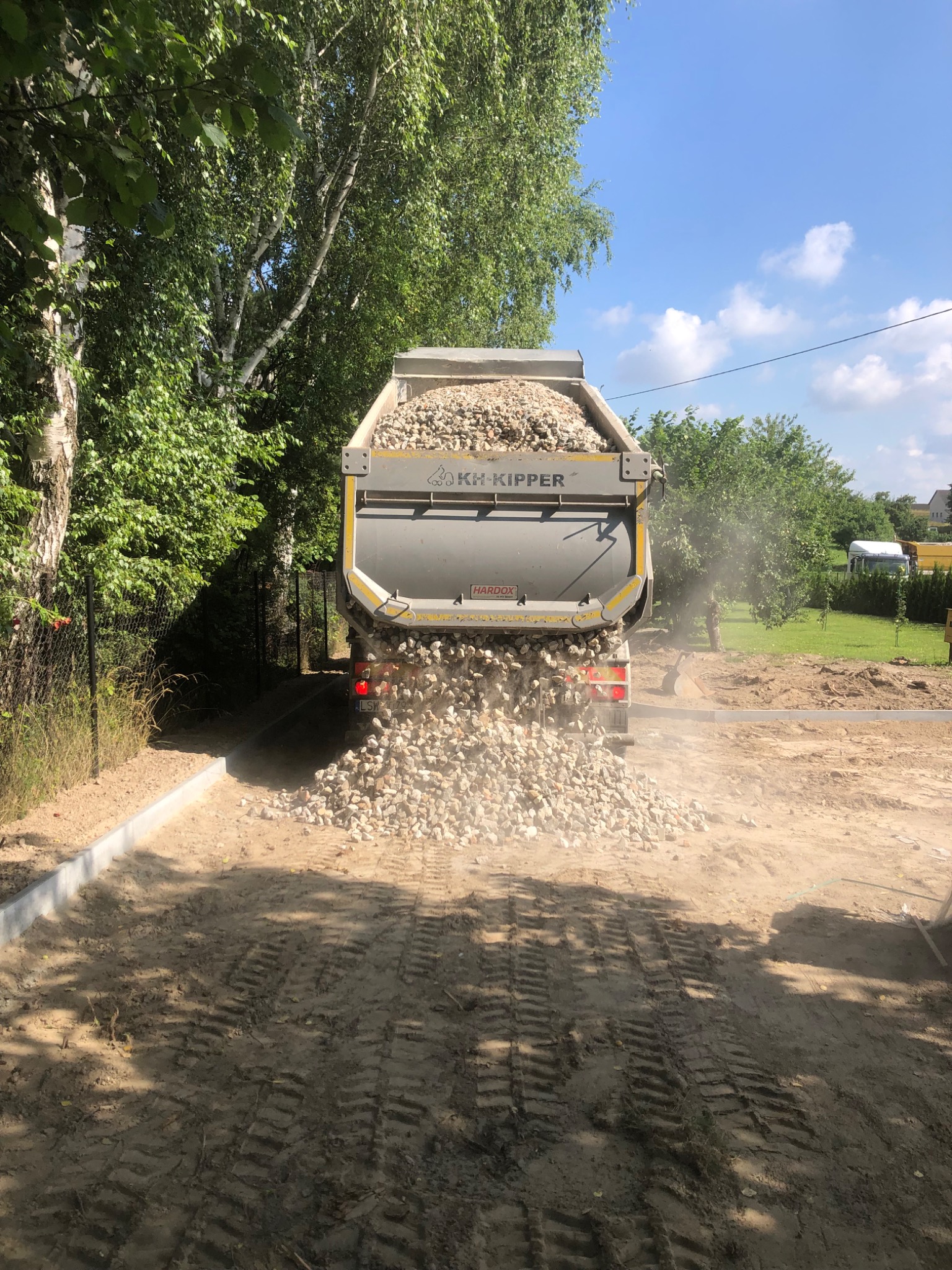 Wywrotka z otwartą skrzynią wysypuje żwir na przygotowany teren z obrzeżami. Widoczne ślady opon i zieleń w tle. Etap utwardzania drogi dojazdowej.