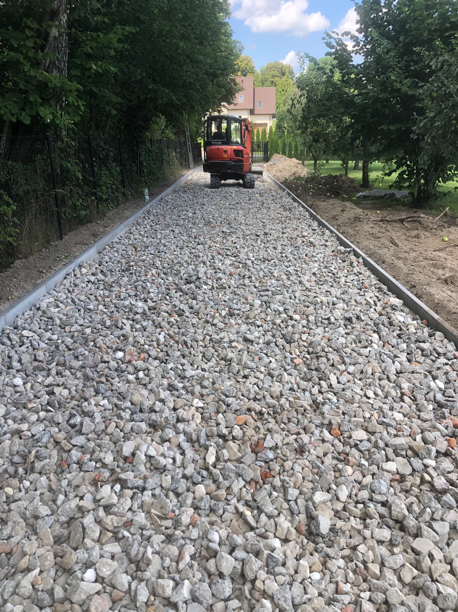 Utwardzona droga z szarym kamieniem i obrzeżami, w tle mała koparka Kubota i zieleń drzew. Widok z perspektywy.