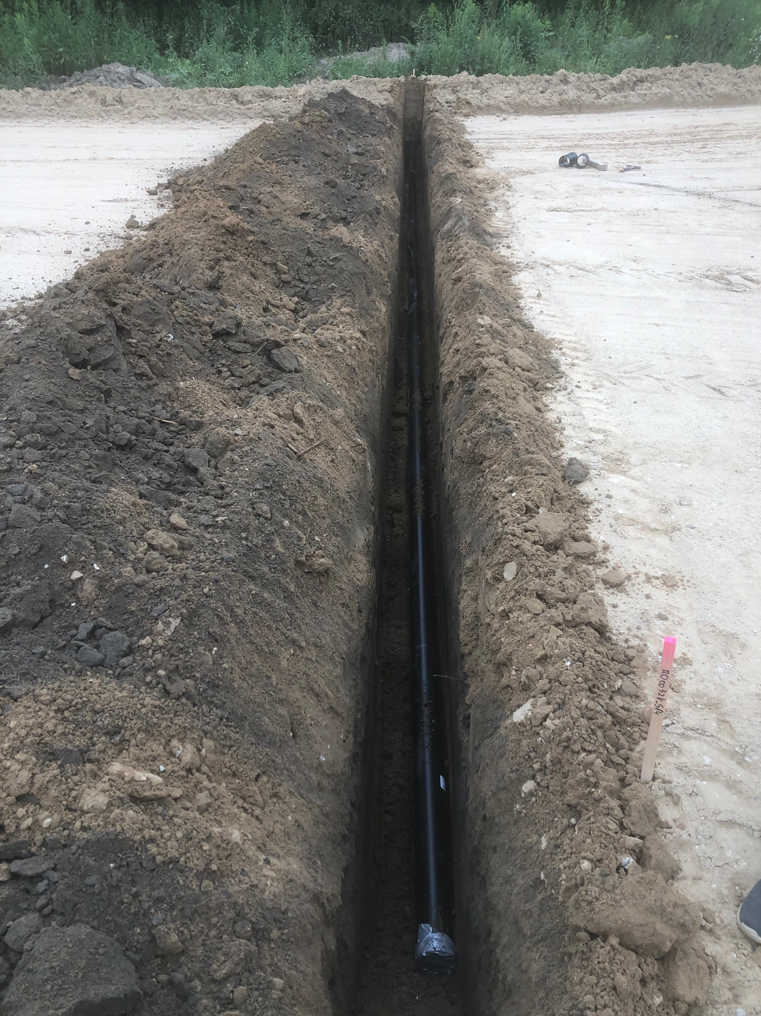 Wąski wykop w ziemi z ułożoną czarną rurą. Widoczne narzędzia i oznaczenia geodezyjne. Świeżo wykopana ziemia.