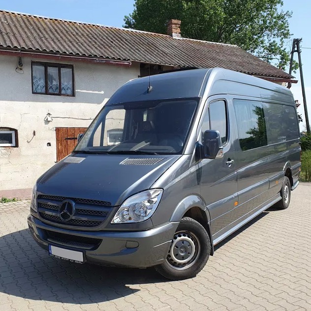 Szary Mercedes Sprinter zaparkowany przed białym budynkiem z brązowymi drzwiami i oknami z białymi firankami, sfotografowany w słoneczny dzień.