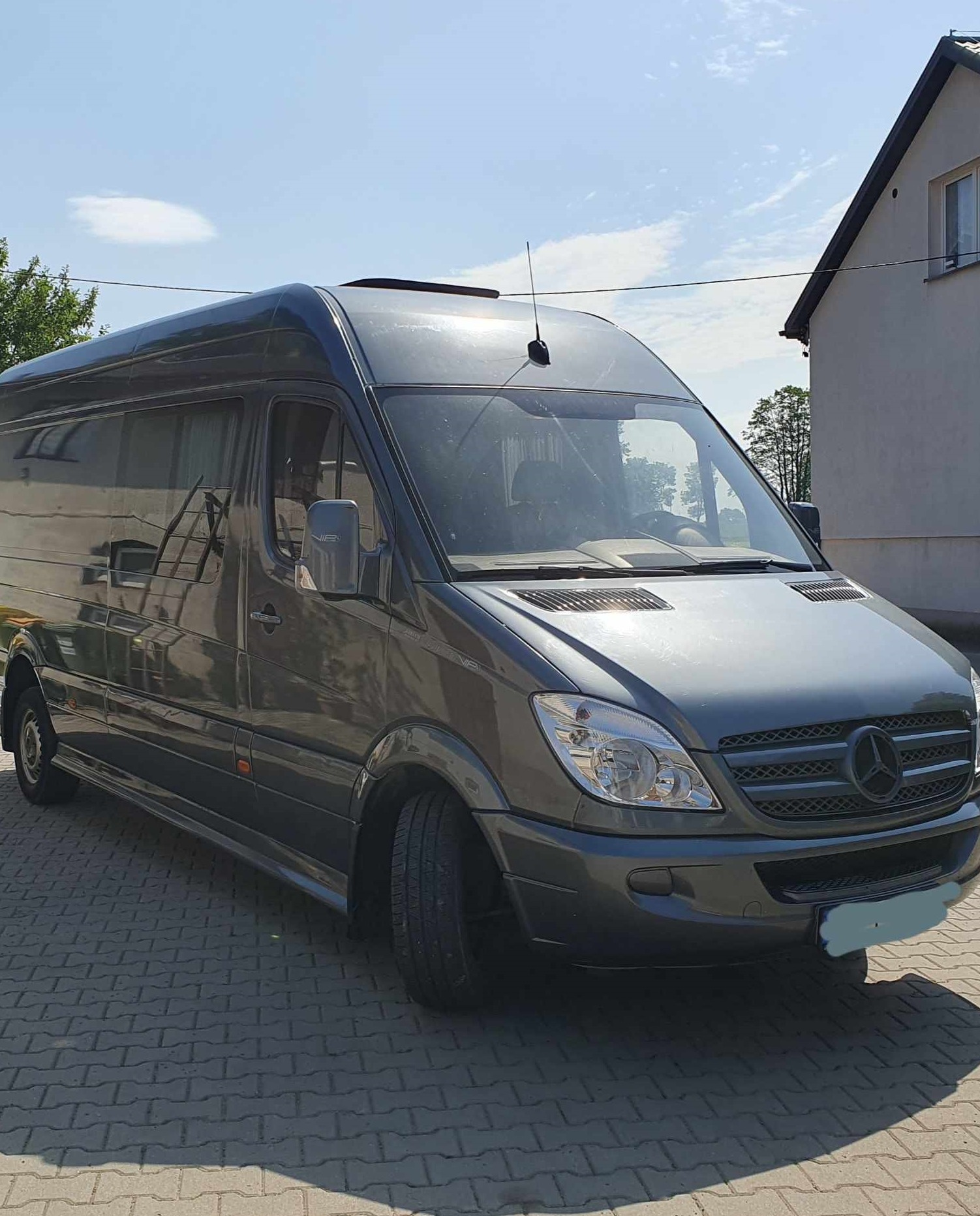 Szary Mercedes-Benz Sprinter z wysokim dachem i anteną na dachu, zaparkowany na brukowanym podjeździe przed budynkiem w słoneczny dzień.