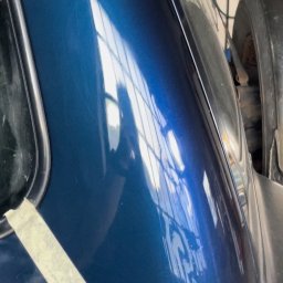 Shine - Błyszczący, niebieski fragment karoserii samochodu z widocznymi zarysowaniami i taśmą maskującą, odbijający jasne okno.