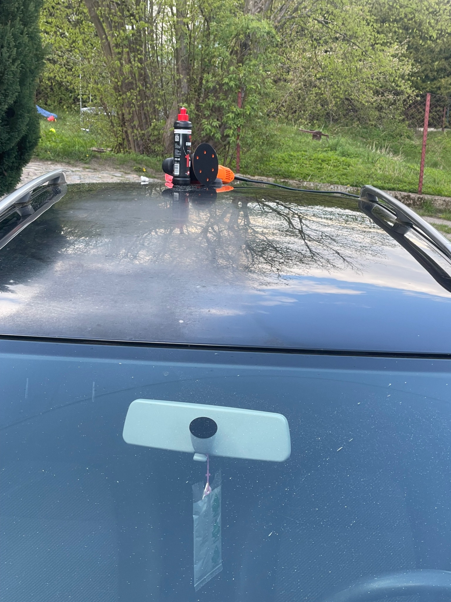 Polerka samochodowa, butelka środka polerskiego i gąbka leżą na dachu czarnego samochodu, odbijającym niebo i drzewa. Widoczna przednia szyba z lusterkiem wstecznym i zawieszką zapachową.