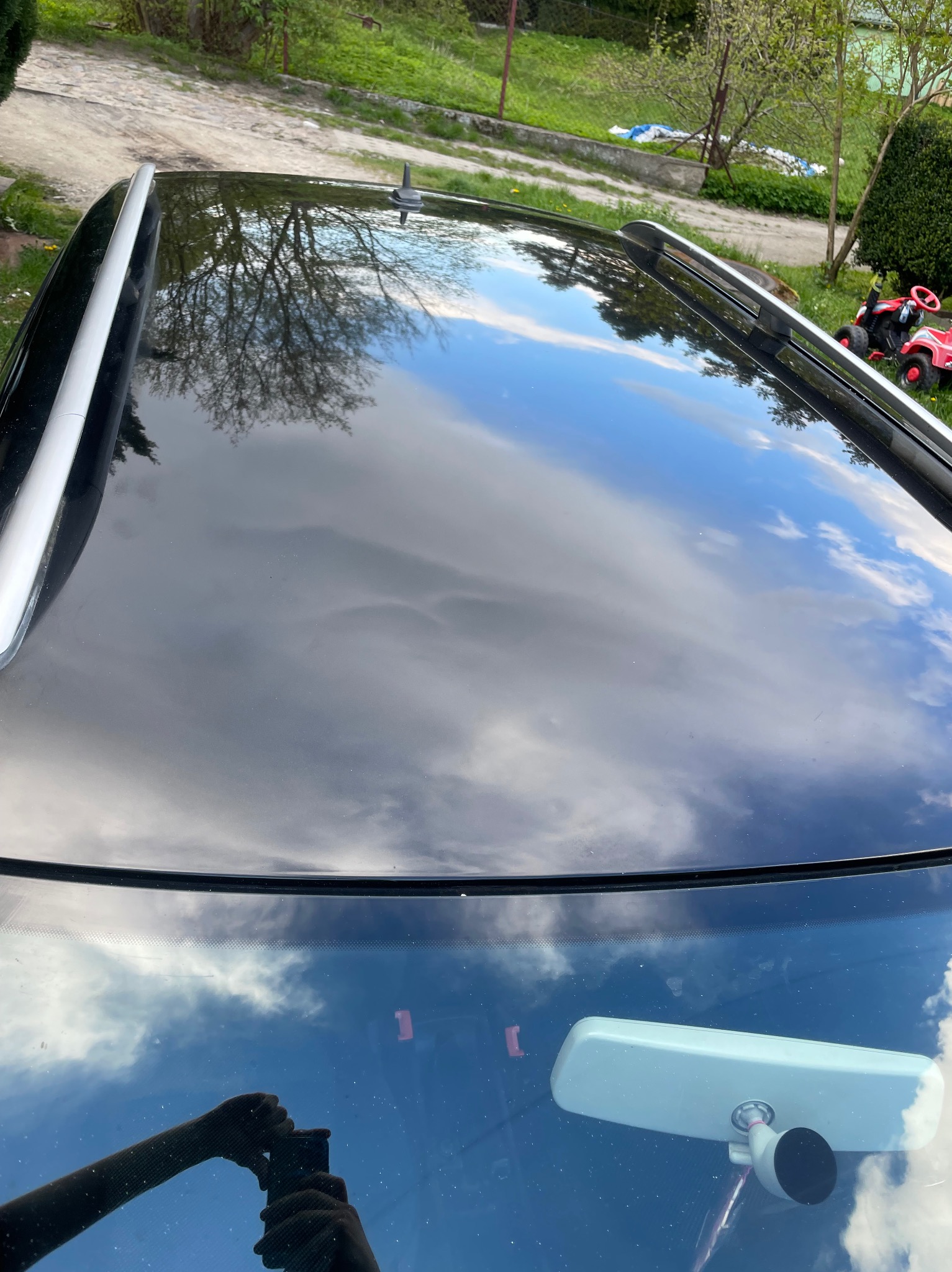 Błyszczący dach czarnego samochodu odbija chmury i drzewa, widoczny przedni panel i lusterko wsteczne, w odbiciu dłonie fotografa.