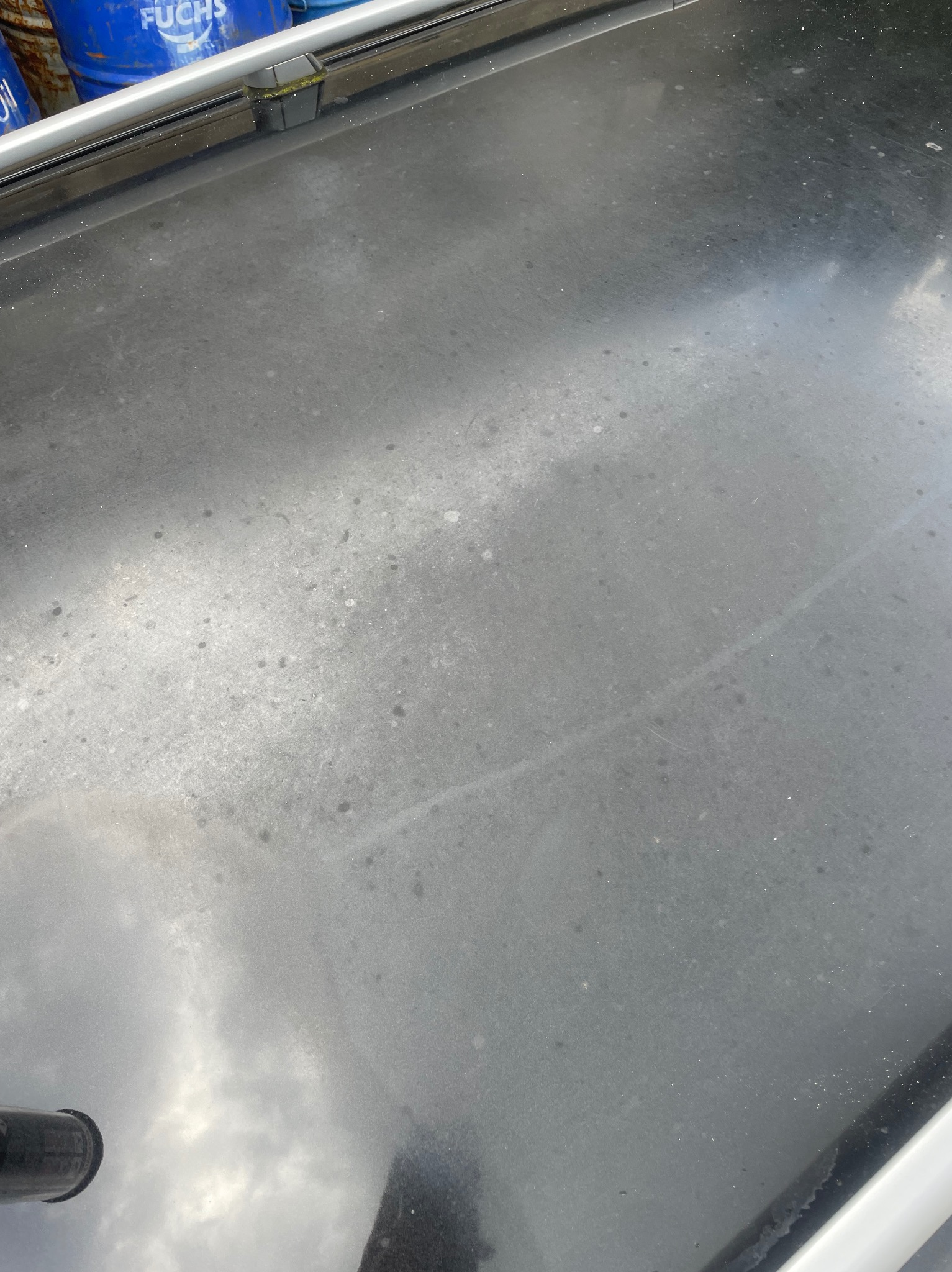 Szara, matowa powierzchnia z widocznymi plamami i smugami, częściowo odbijająca niebo, z fragmentem niebieskich beczek w tle.