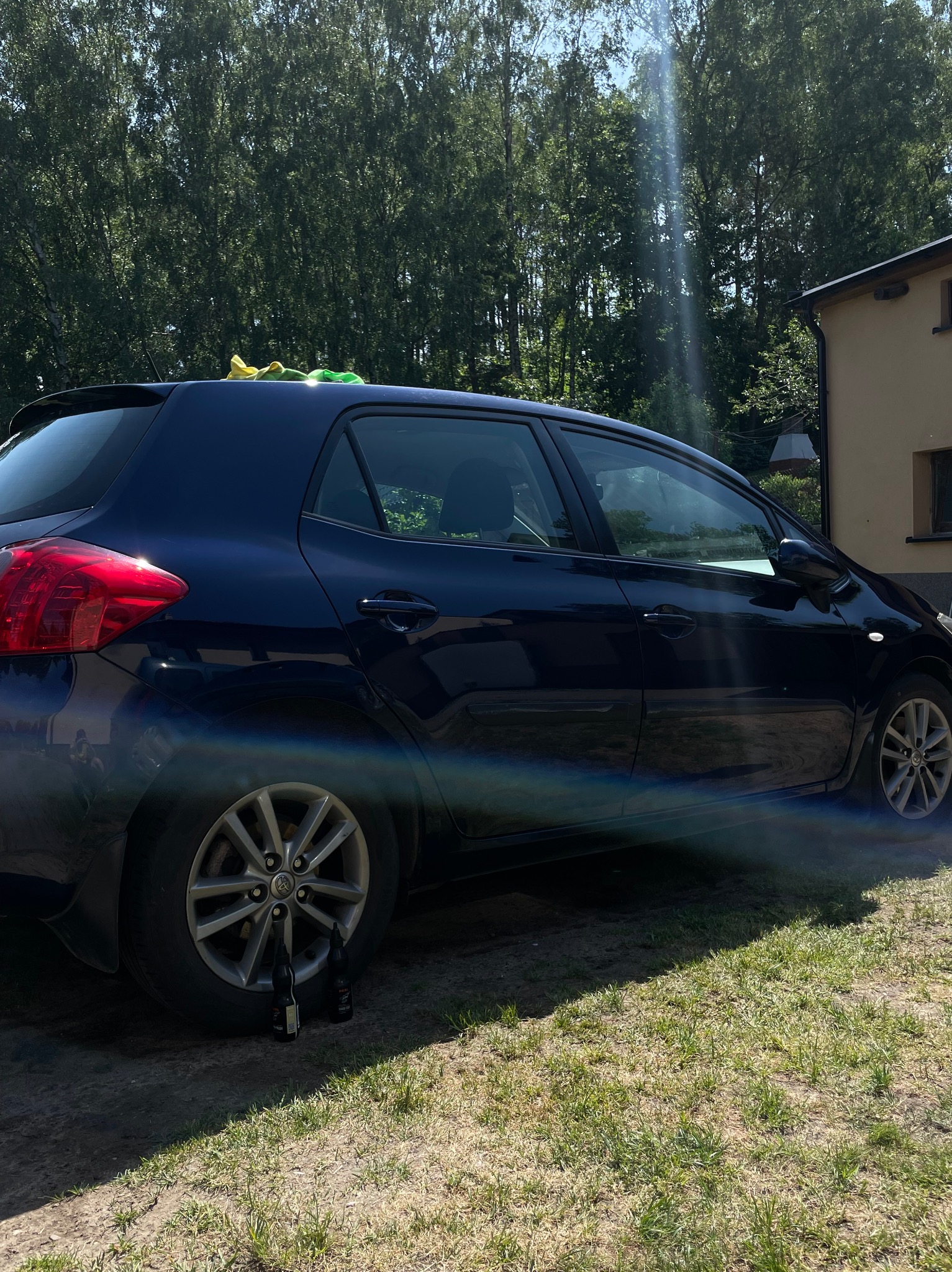 Granatowy samochód z zielonymi i żółtymi ściereczkami na dachu, stojący na trawie obok budynku, z dwoma butelkami pod kołem.